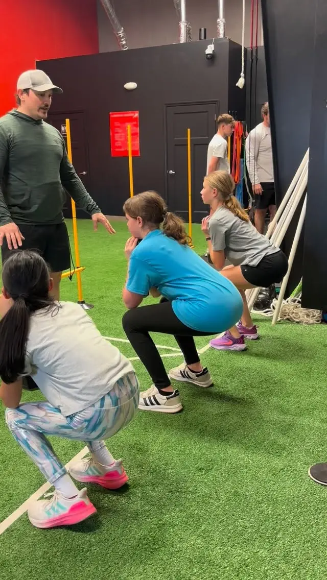 Coach leading three girls through synchronized squats on the green turf
