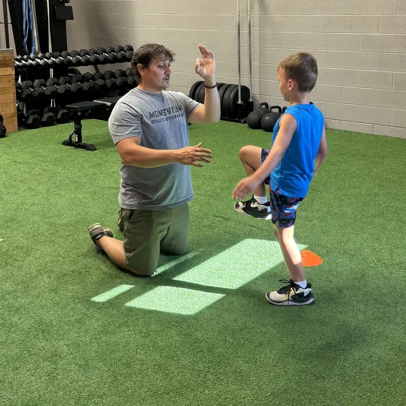 A Momentum coach kneeling on the turf teaching knee drive form one-on-one with a young Jump Start athlete