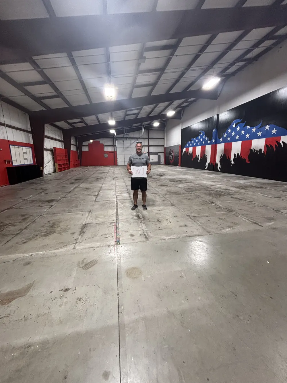 Coach Ryan Cheek standing alone in the middle of the empty warehouse floor holding a handwritten Day 1 sign