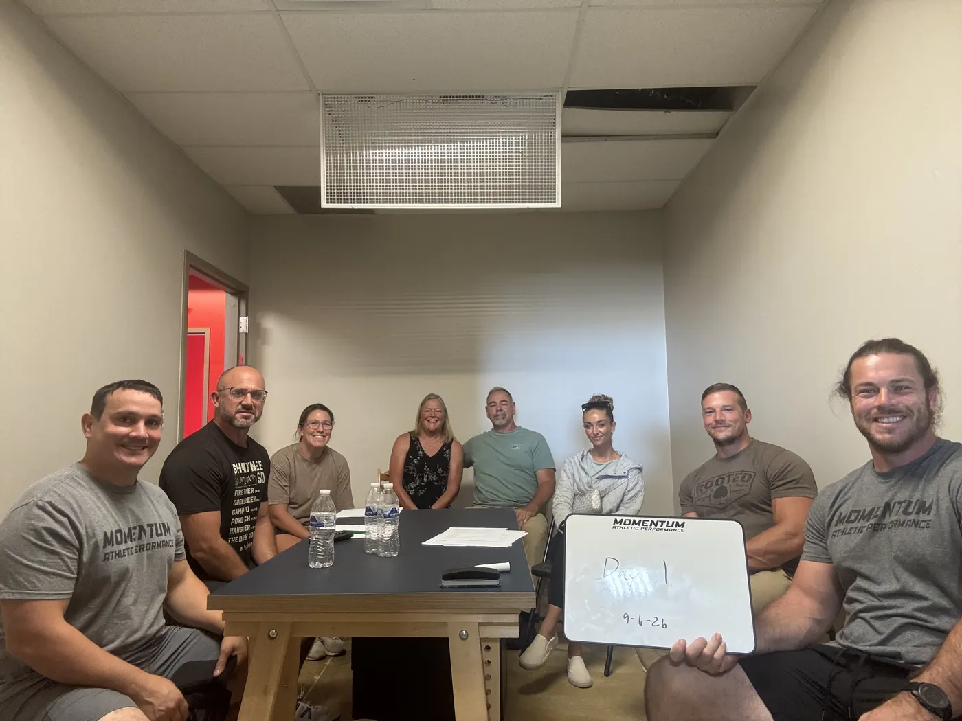 Eight members of the Momentum team seated around a table in the new building's meeting room, one holding a small whiteboard